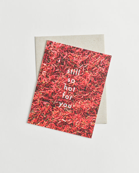 Photo greeting card of a bed of red chili peppers and white words “still so hot for you”.
